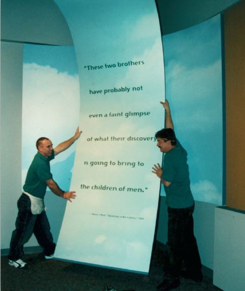 Two workers install an interpretive piece with a quote from Amos Ives Root